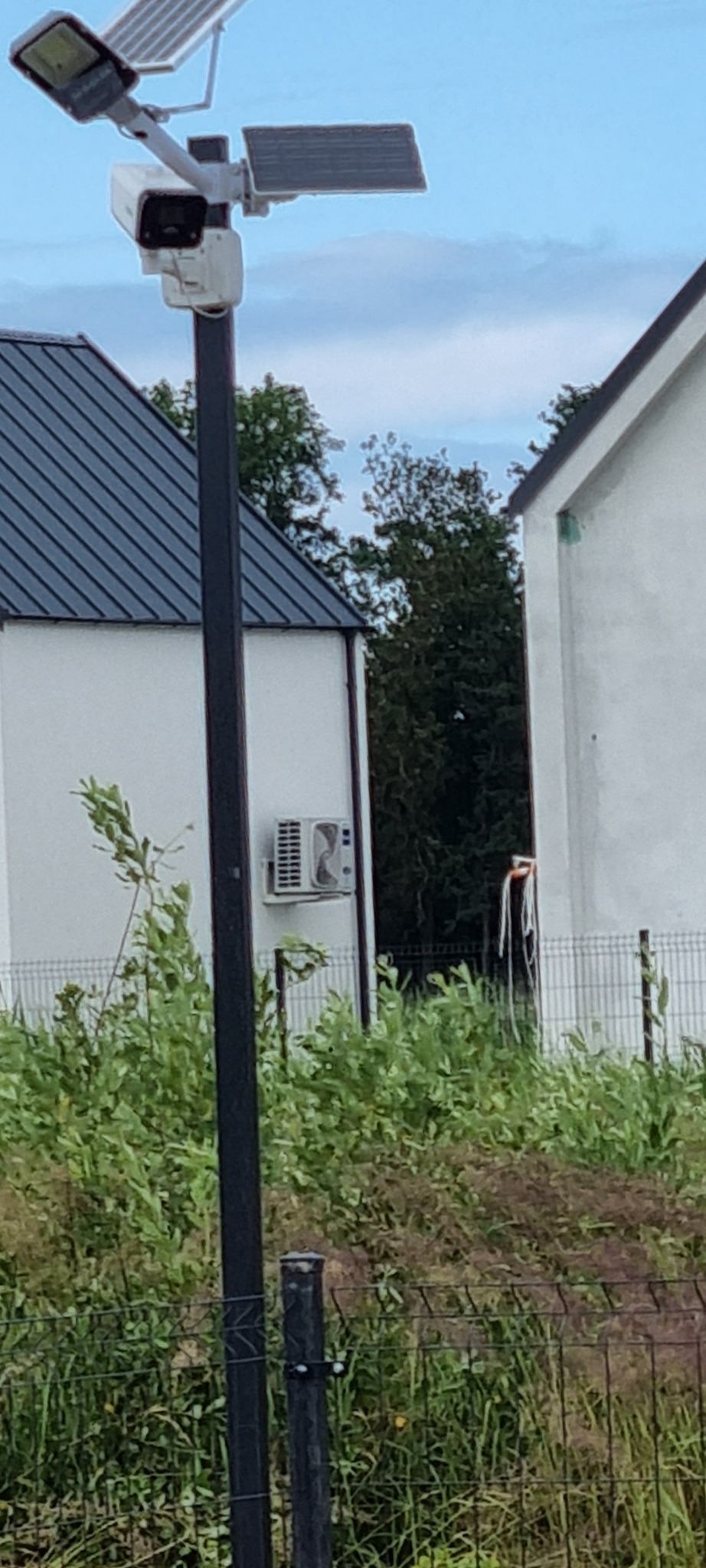 Kamera monitoringu z panelem solarnym na słupie, widok zza ogrodzenia. W tle budynki mieszkalne i zieleń. Widoczne elementy systemu monitoringu.