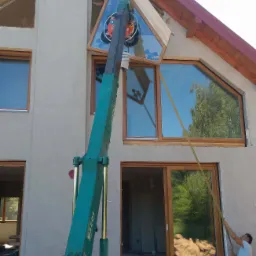 Montaż okna o nietypowym trójkątnym kształcie za pomocą dźwigu na placu budowy domu jednorodzinnego.