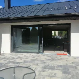 Nowoczesny dom z dużym, przesuwnym oknem tarasowym w grafitowej ramie, widok z zewnątrz na częściowo wykończone wnętrze i taras z szarej kostki brukowej.