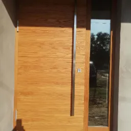 Nowoczesne drzwi wejściowe z drewna z długim, pionowym uchwytem ze stali nierdzewnej i bocznym panelem szklanym, odbijającym otoczenie zewnętrzne.