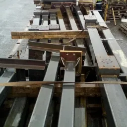 Stalowe profile i ramy, prawdopodobnie do konstrukcji bram, ułożone na palecie w deszczowy dzień na placu budowy, w tle palety i budynki.