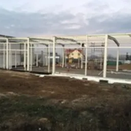 Konstrukcja stalowa hali w trakcie budowy, widoczne białe elementy szkieletu, w tle dom jednorodzinny i niebo z lekkim zachmurzeniem.