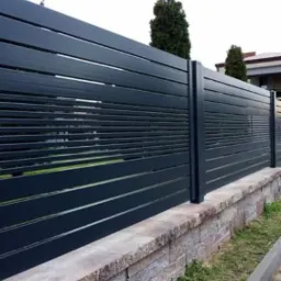 Nowoczesna, pozioma, metalowa brama wjazdowa w kolorze antracytowym, zamontowana na kamiennym murku, z widocznymi szczelinami między panelami i roślinnością w tle.