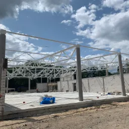 Konstrukcja stalowa hali w trakcie budowy, widoczne betonowe słupy, białe kratownice dachu, ściany z bloczków oraz pracownik w tle.