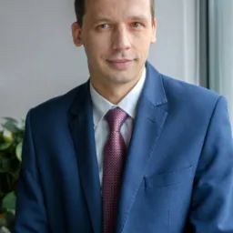 Portret mężczyzny w niebieskim garniturze i bordowym krawacie w kropki, stojącego na tle jasnej ściany i rośliny doniczkowej.