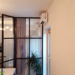 Wnętrze domu z klimatyzatorem na ścianie, widoczne przeszklenie w czarnej ramie, zasłona, roślina doniczkowa i grzejnik podłogowy.