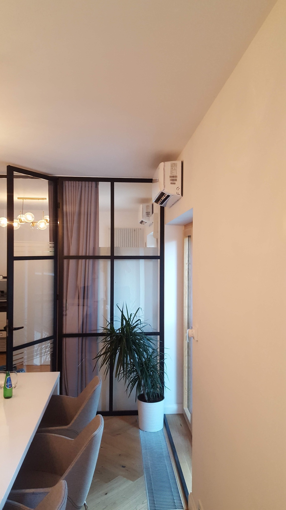 Wnętrze domu z klimatyzatorem na ścianie, widoczne przeszklenie w czarnej ramie, zasłona, roślina doniczkowa i grzejnik podłogowy.