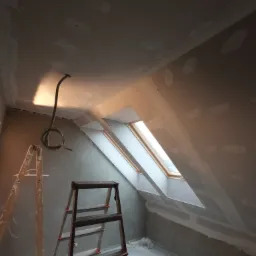Poddasze w trakcie prac wykończeniowych: widoczne szpachlowane łączenia płyt gipsowo-kartonowych, okno dachowe, dwie drabiny i zwisający przewód elektryczny.