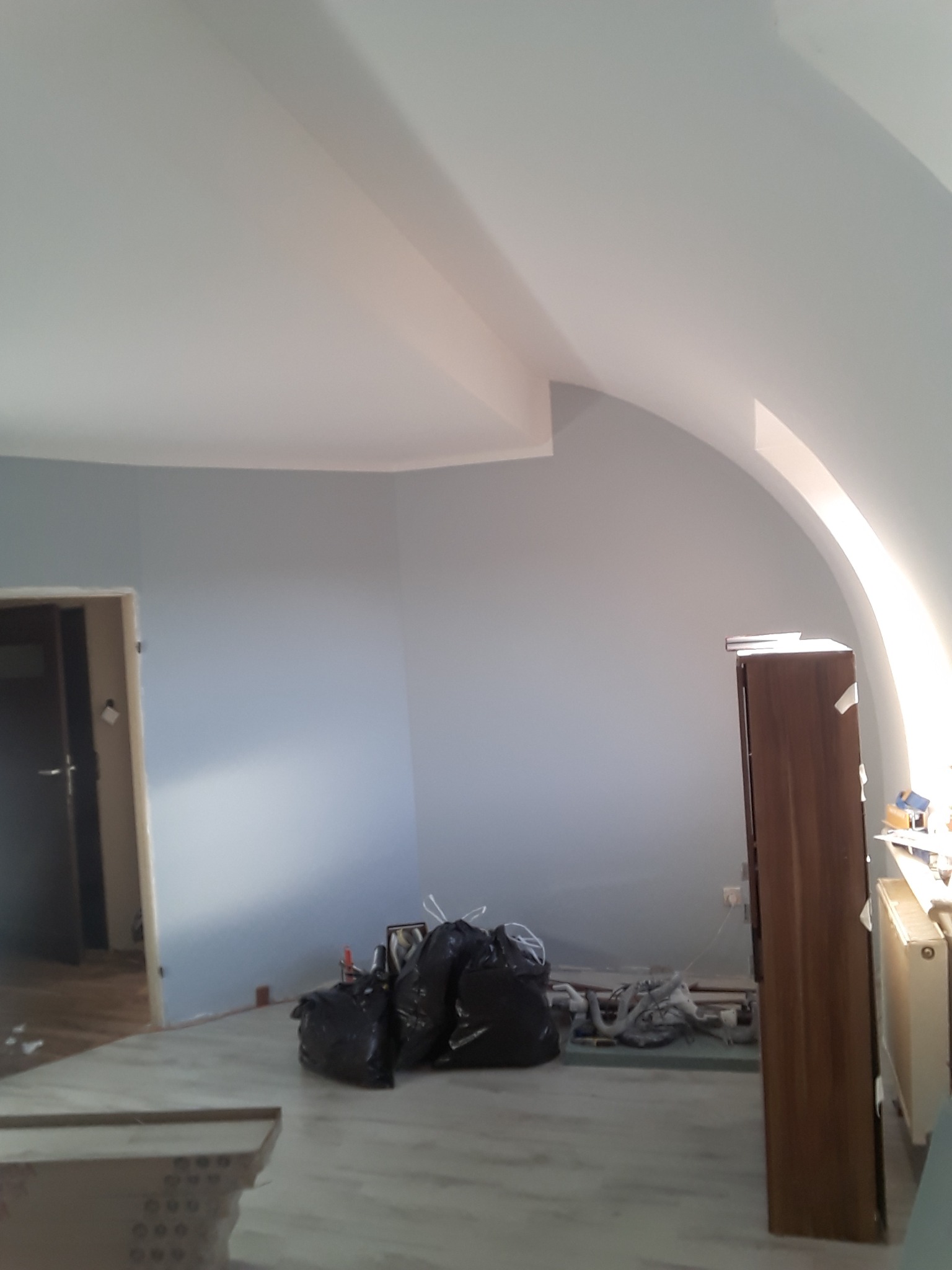Pokój na poddaszu w trakcie odświeżania: świeżo pomalowane na szaro ściany, biały sufit ze skosami, zabezpieczone folią podłogi, worki na śmieci i narzędzia, widoczny fragment drewnianej szafy.
