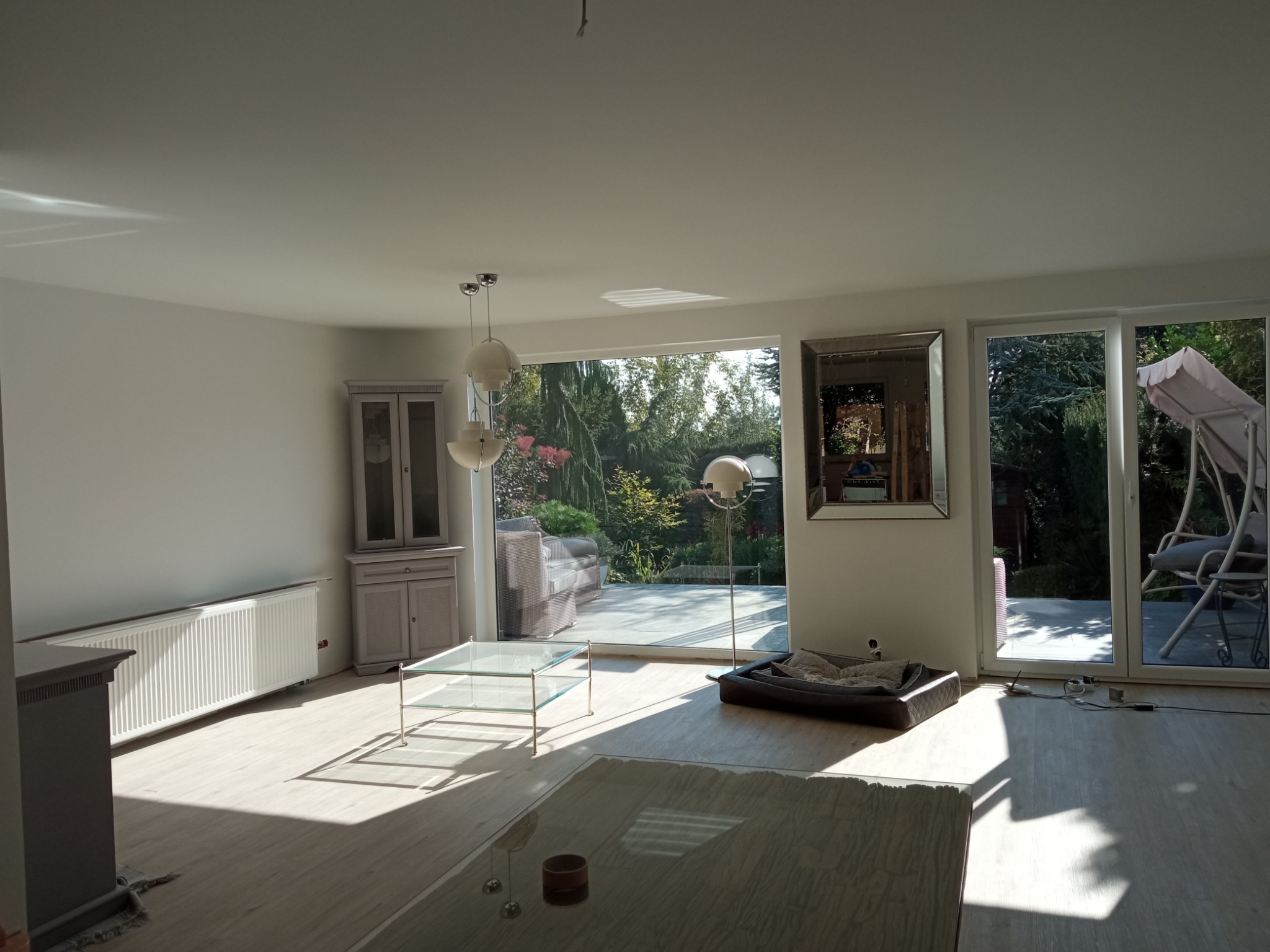 Jasne, przestronne wnętrze salonu po remoncie, z widokiem na ogród przez duże okna tarasowe, świeżo położona podłoga, pomalowane ściany na biało, nowoczesne oświetlenie i meble.