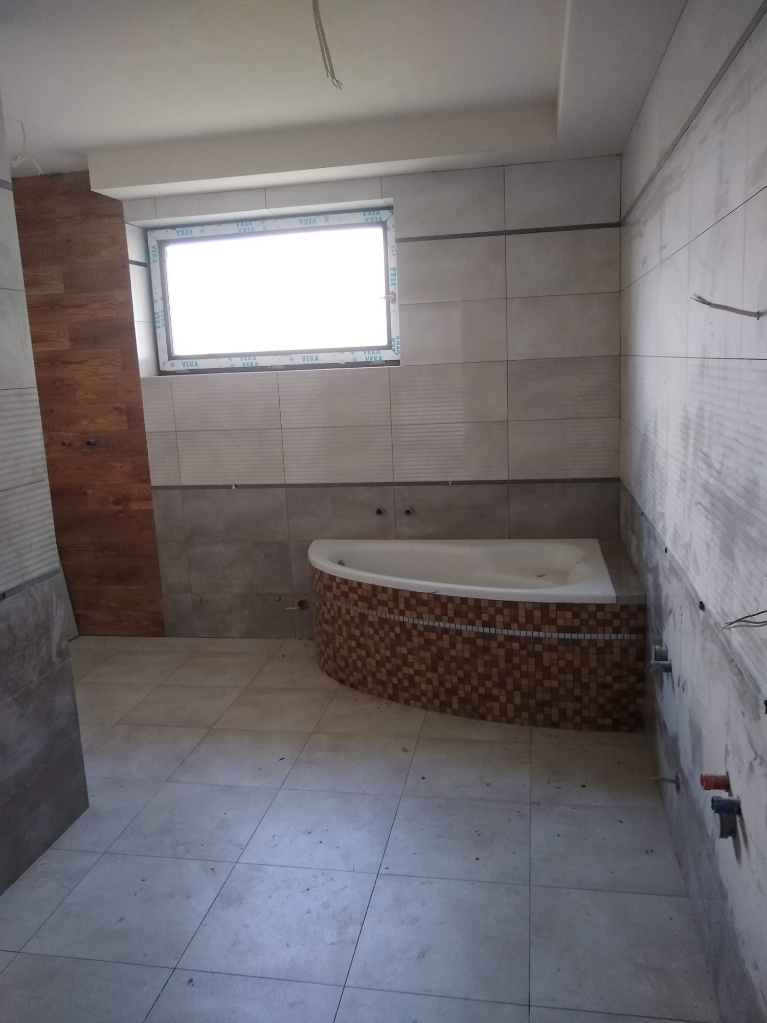 Łazienka w trakcie remontu: ułożone płytki na podłodze i częściowo na ścianach, obudowana mozaiką wanna narożna, widoczne instalacje hydrauliczne i elektryczne, okno z widocznym logotypem producenta.
