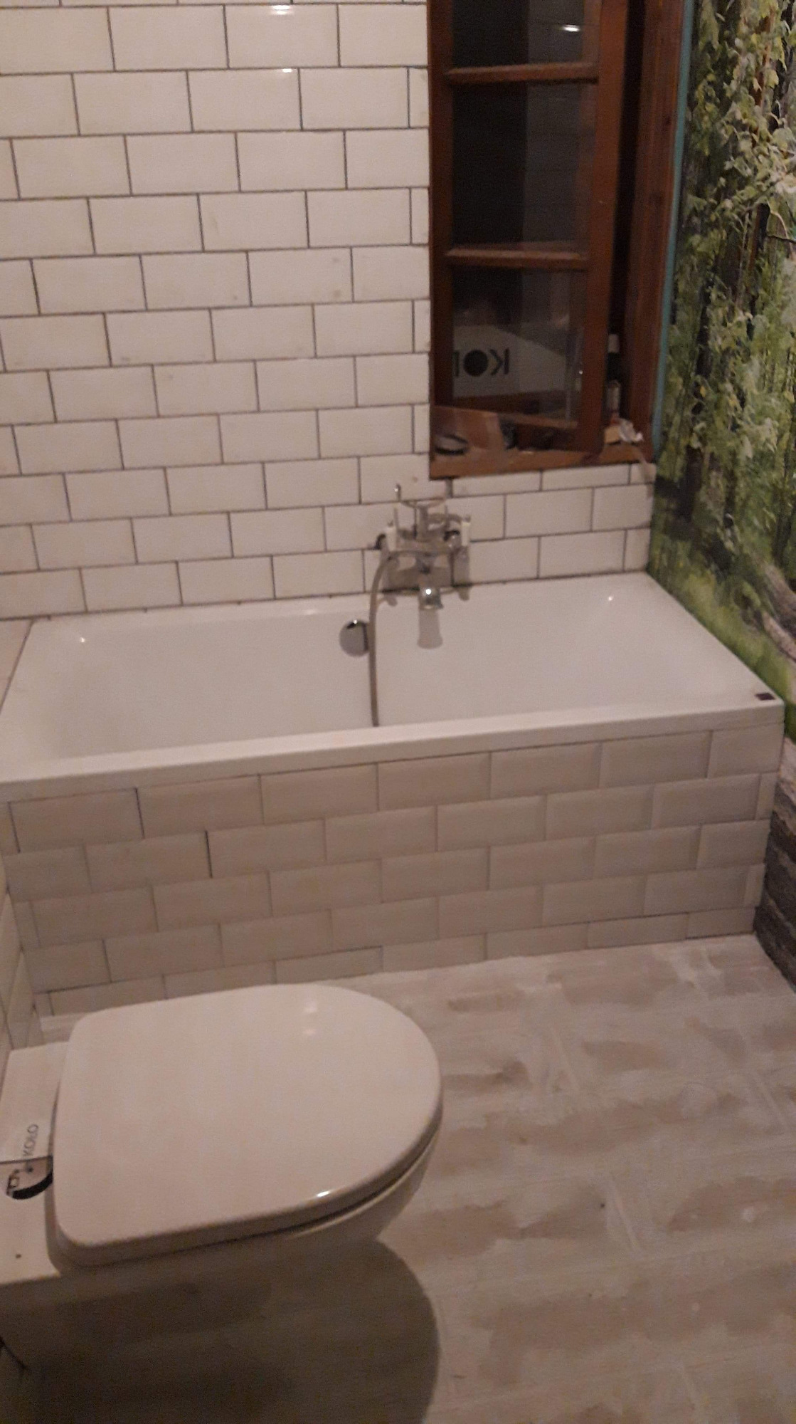 Wykończona łazienka z białą wanną obudowaną płytkami, toaletą marki Koło oraz drewnianą ramą okienną z widocznym fragmentem plakatu w tle i fototapetą z motywem leśnym po prawej stronie.