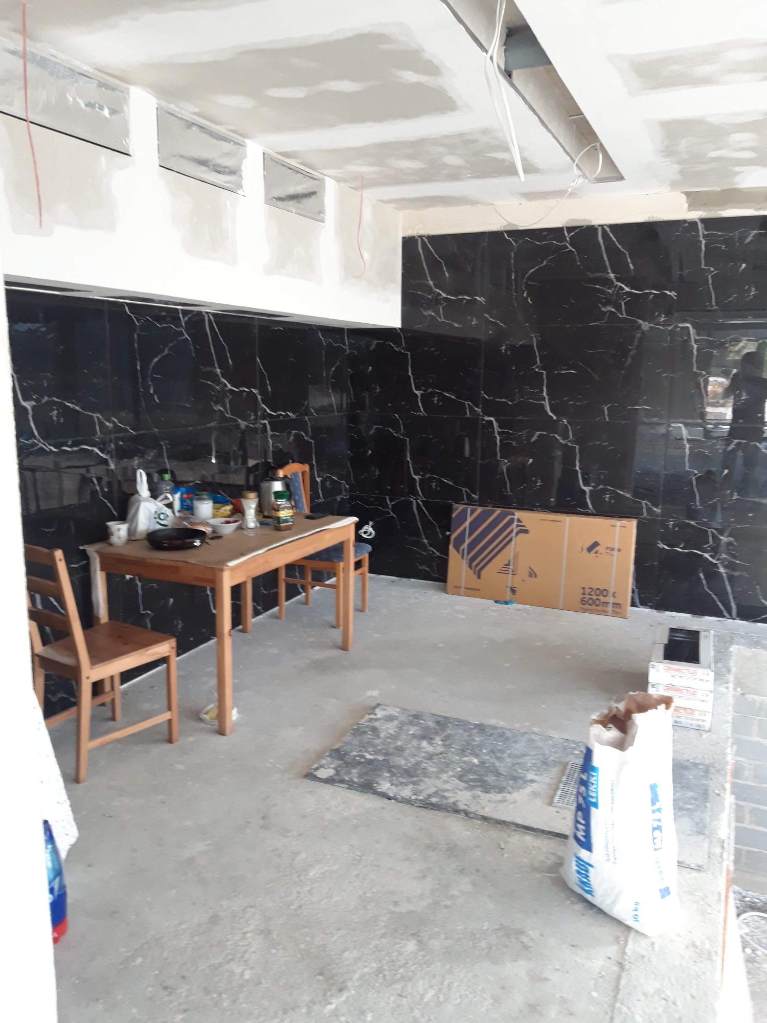 Wnętrze w trakcie prac remontowych: czarne płytki imitujące marmur na ścianach, surowy sufit z płytami, stół z krzesłami i karton po płytkach na podłodze.
