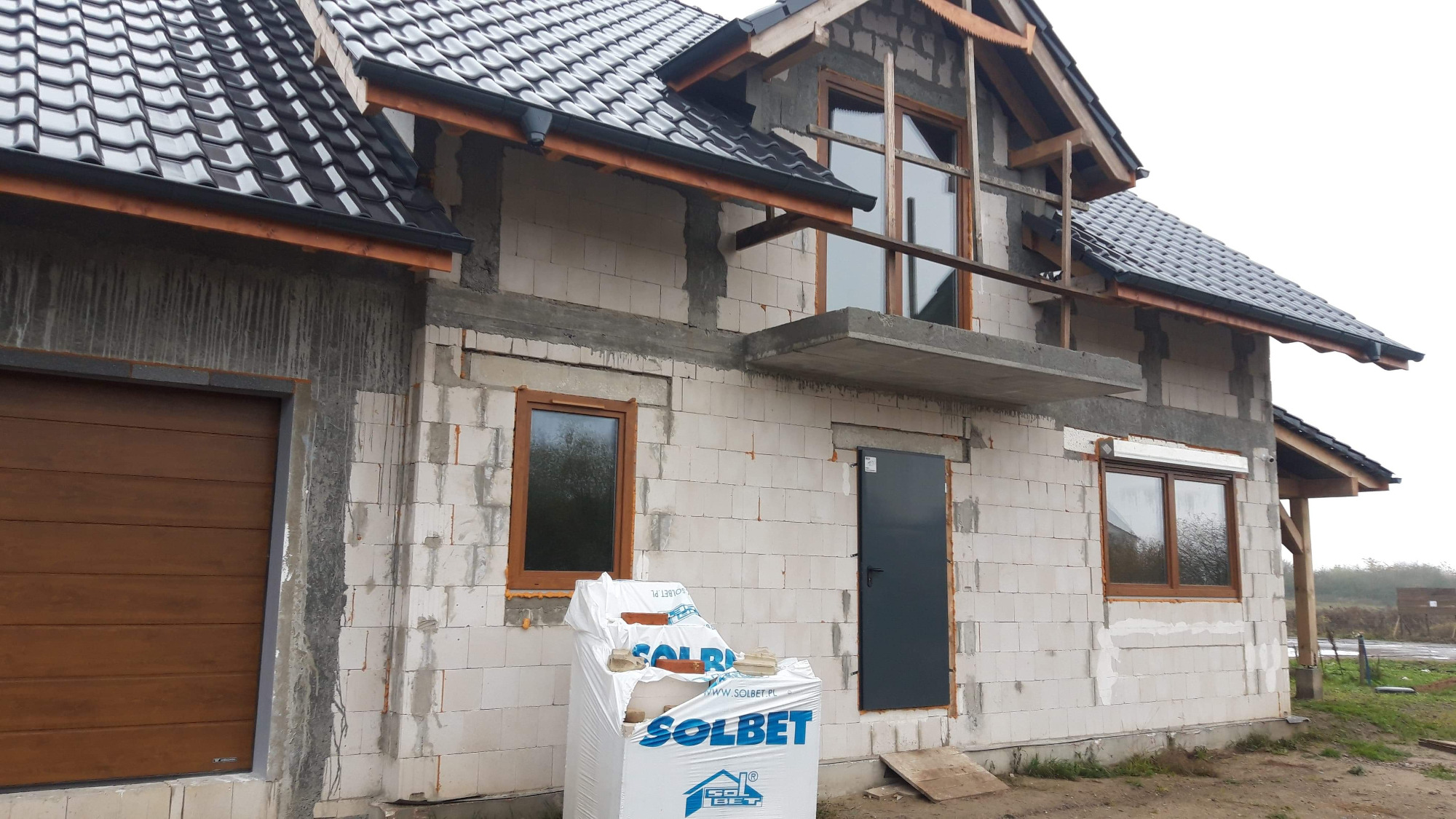 Budynek w trakcie budowy z widocznymi białymi bloczkami Solbet, brązowymi oknami i drzwiami, oraz częściowo pokrytym dachem z ciemnej dachówki.