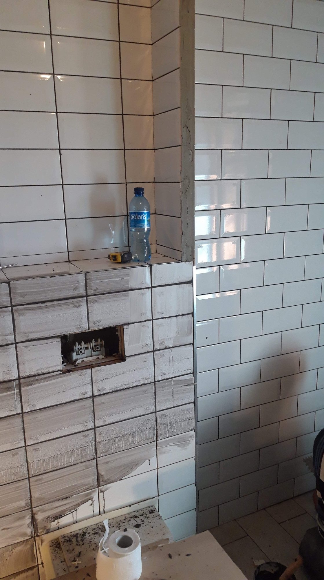 Układanie białych, prostokątnych płytek na ścianie w łazience, widoczny otwór na gniazdko w trakcie prac, butelka wody i miarka na półce, częściowo zabrudzone fugi.