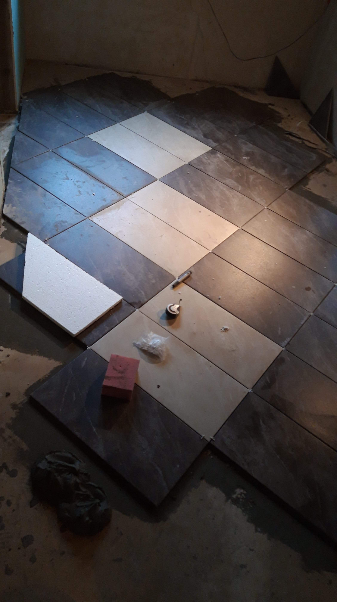 Układanie płytek podłogowych w romby, z użyciem płytek w dwóch odcieniach szarości, widoczny fragment białej płyty styropianowej, szpachelka, miarka, gąbka i zaprawa.