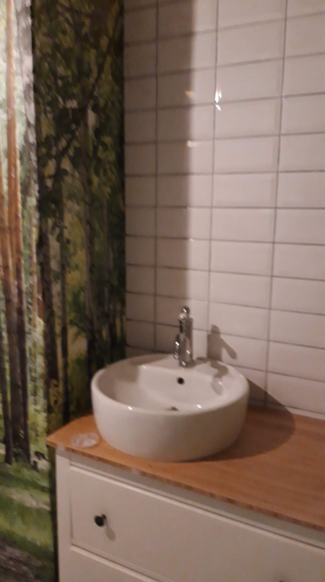 Okrągła umywalka nablatowa z chromowaną baterią na blacie z jasnego drewna, białe płytki na ścianie i tapeta z motywem leśnym w tle.