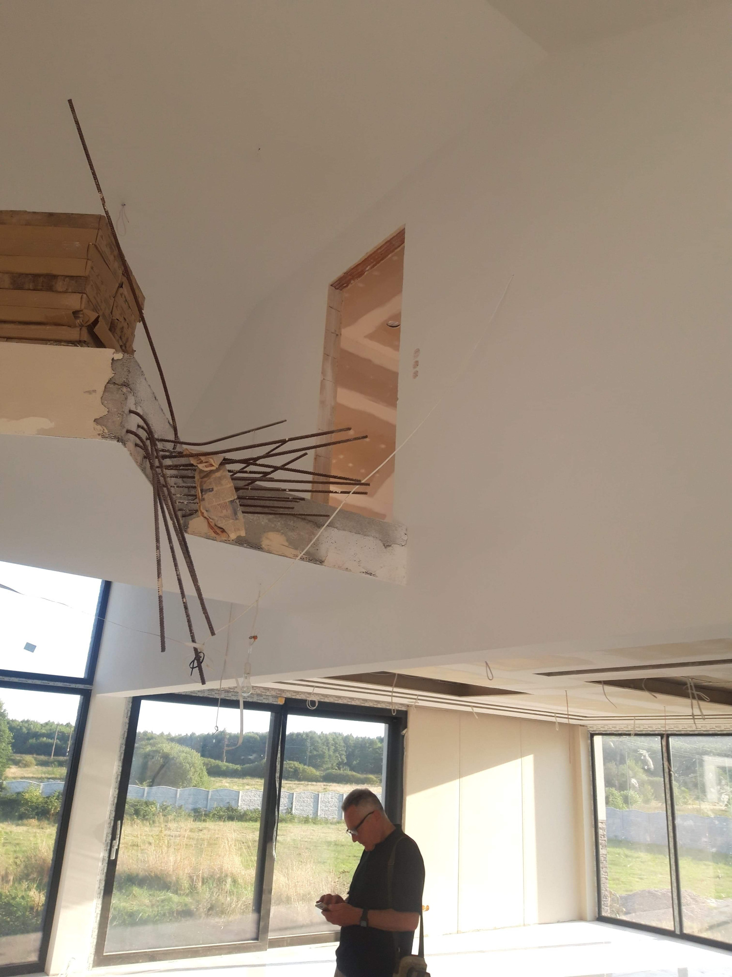 Wnętrze w trakcie remontu z widocznym zbrojeniem wystającym ze stropu, otwór okienny w ścianie i mężczyzna z telefonem w tle, duże okna wychodzące na zewnątrz.
