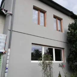 Świeżo otynkowana elewacja dwukondygnacyjnego budynku z nowymi oknami, widoczne elementy rusztowania i kwiaty w ogrodzie.