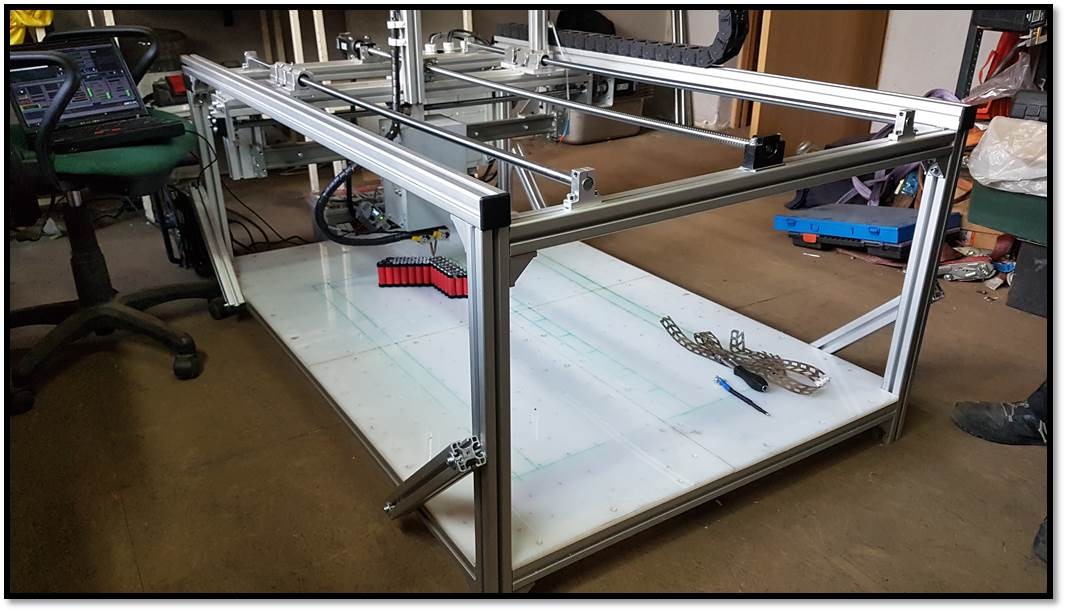 Rama z profili aluminiowych, prawdopodobnie maszyny CNC, z widocznymi elementami sterującymi, laptopem i akcesoriami na blacie roboczym z siatką linii.