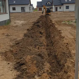 Wąski wykop pod instalacje na piaszczystym terenie budowy, w tle szereg identycznych domów jednorodzinnych, mała koparka w trakcie pracy.