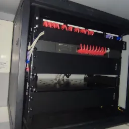 Otwarta szafa rack z zamontowanymi urządzeniami sieciowymi, widoczne kable i listwa z czerwonymi uchwytami, obok biały panel z napisem.