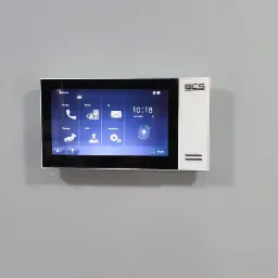 Biały panel dotykowy wideodomofonu BCS zamontowany na szarej ścianie, wyświetlający menu główne z ikonami połączeń, wiadomości i ustawień.