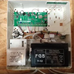Wnętrze białej obudowy centrali alarmowej z widoczną płytą główną Satel PERFECTA 16, transformatorem Pulsar AWT150 i akumulatorem FGB 7-12, zamontowane na płycie OSB.