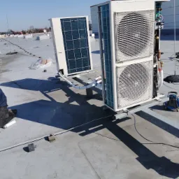 Instalacja zewnętrznych jednostek klimatyzacji na płaskim dachu budynku komercyjnego. Pracownik w kombinezonie podłącza przewody, widoczne inne jednostki w tle.