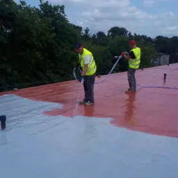 Dwóch pracowników w kamizelkach odblaskowych natryskuje farbę na płaski dach budynku, część dachu pomalowana na szaro, a część na czerwono, widoczne przewody i dysze natryskowe.