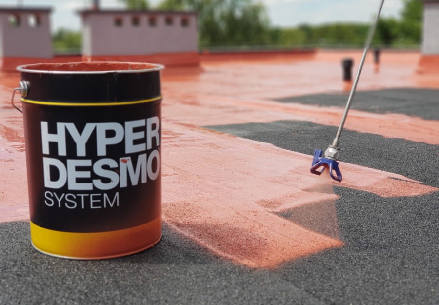 Puszka farby Hyper Desmo System stoi obok pistoletu natryskowego na dachu z częściowo pomalowaną powierzchnią na pomarańczowo, w tle kominy i zieleń.