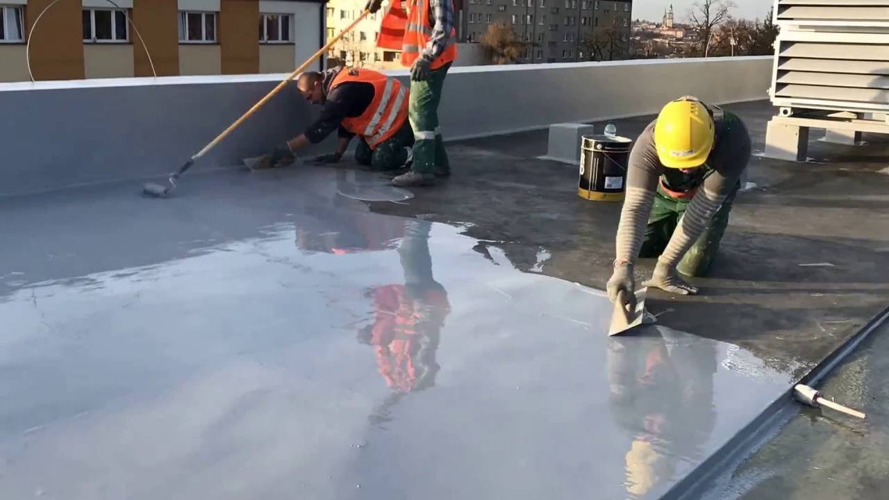 Praca na dachu płaskim: trzech robotników w kamizelkach odblaskowych i kaskach nanosi szarą powłokę hydroizolacyjną za pomocą wałka i szpachelek, widoczny budynek w tle.