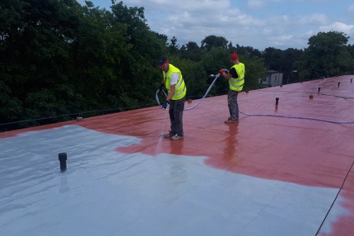 Dwóch pracowników w kamizelkach odblaskowych natryskuje farbę na płaski dach budynku, część dachu pomalowana na szaro, a część na czerwono, widoczne przewody i dysze natryskowe.