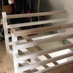 MMK Marcel Pieczka - Biała, drewniana konstrukcja antresoli w trakcie montażu, widoczne elementy stelaża i podłogi, ubrania wiszące w tle.