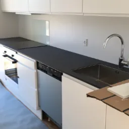 Minimalistyczna kuchnia z białymi szafkami i blatem w kolorze antracytowym, widoczny zlew z chromowaną baterią i płyta indukcyjna, surowy betonowy odcień podłogi w tle.