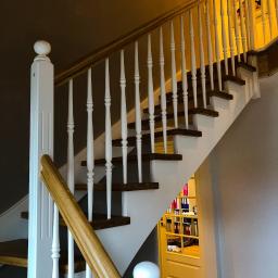 MMK Marcel Pieczka - Klasyczne schody drewniane z białymi balustradami i brązowymi stopniami, widok z dołu, w tle drzwi do pokoju z regałem pełnym segregatorów.