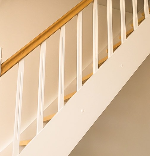 Fragment białej balustrady schodowej z drewnianym poręczą, oświetlonej ciepłym światłem.