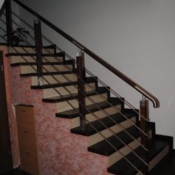 ESLA PRACOWNIA DREWNA I STALI Sp. zo.o. - Wewnętrzne schody z ciemnego drewna i beżowych podstopnic, zabezpieczone metalową balustradą z drewnianą poręczą, widok z dołu na tle ściany z dekoracyjnym tynkiem.