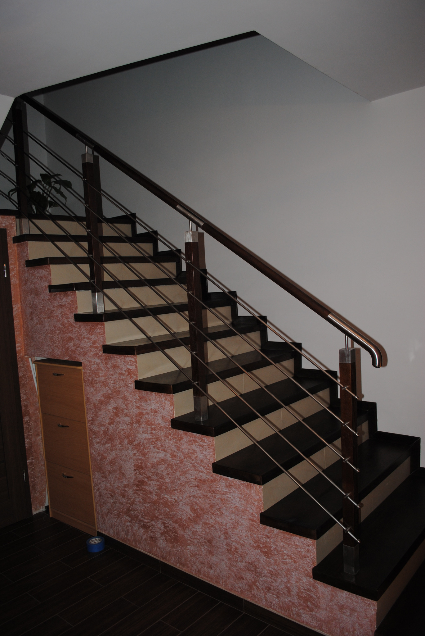 Wewnętrzne schody z ciemnego drewna i beżowych podstopnic, zabezpieczone metalową balustradą z drewnianą poręczą, widok z dołu na tle ściany z dekoracyjnym tynkiem.