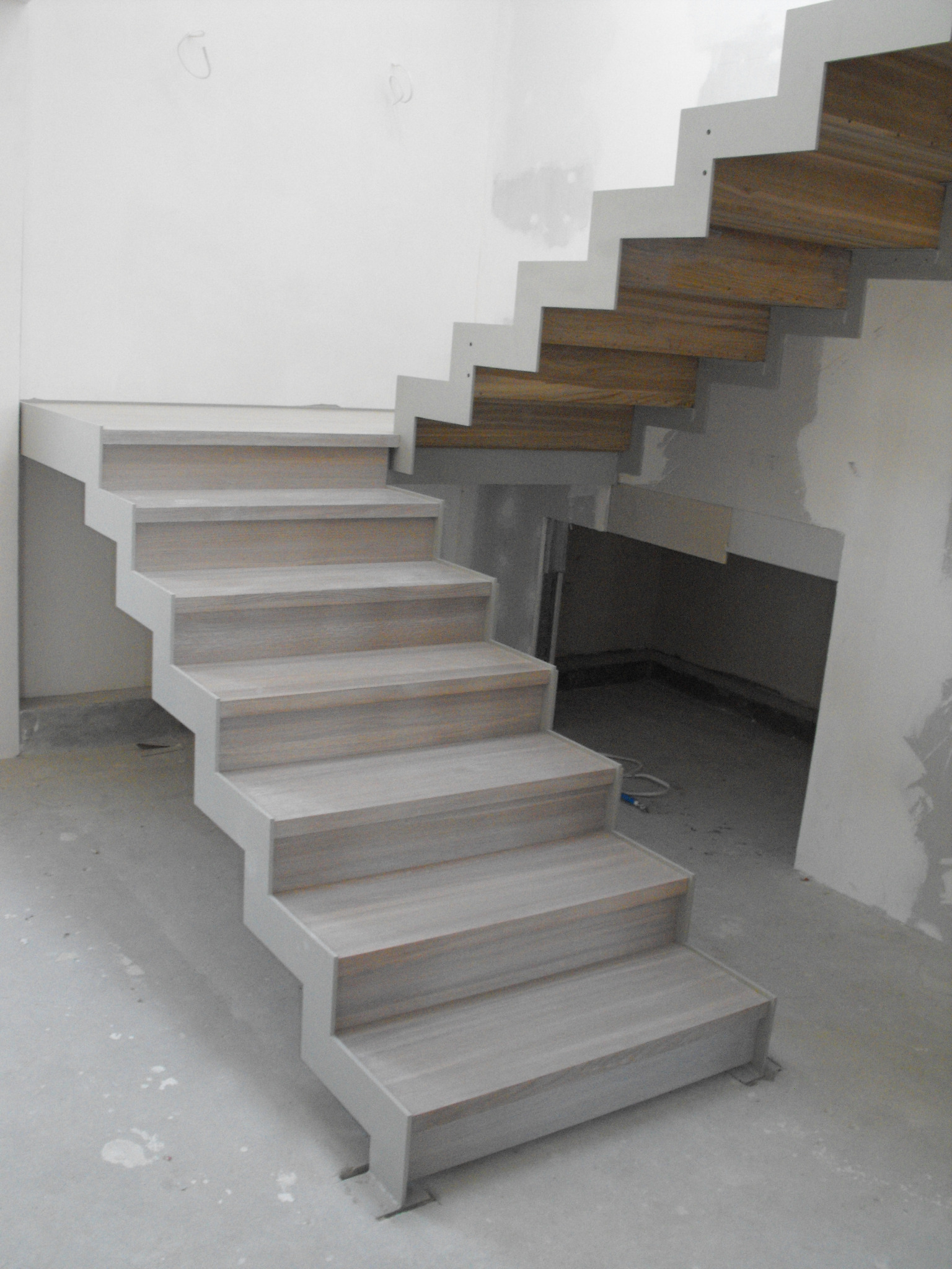 Schody wewnętrzne z drewnianymi stopniami i szarą metalową konstrukcją w surowym wnętrzu podczas remontu.