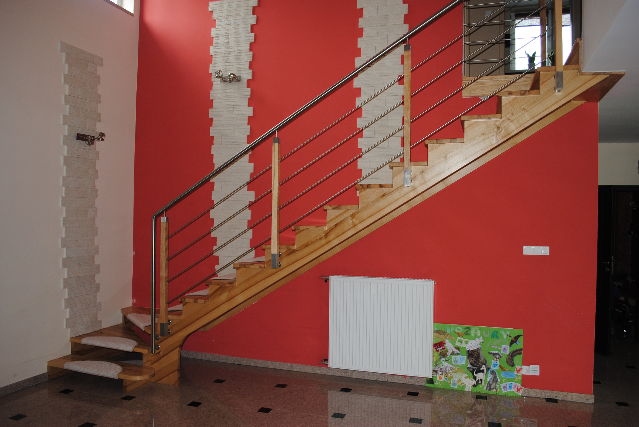 Jasne drewniane schody z nierdzewną balustradą w nowoczesnym wnętrzu z czerwoną ścianą i dekoracyjnymi panelami z kamienia.