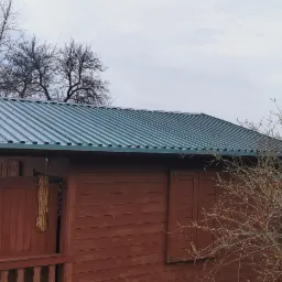 Czerwona drewniana altana z zielonym dachem z blachy trapezowej, widoczna z boku, otoczona drzewami bez liści i krzewami z pąkami.