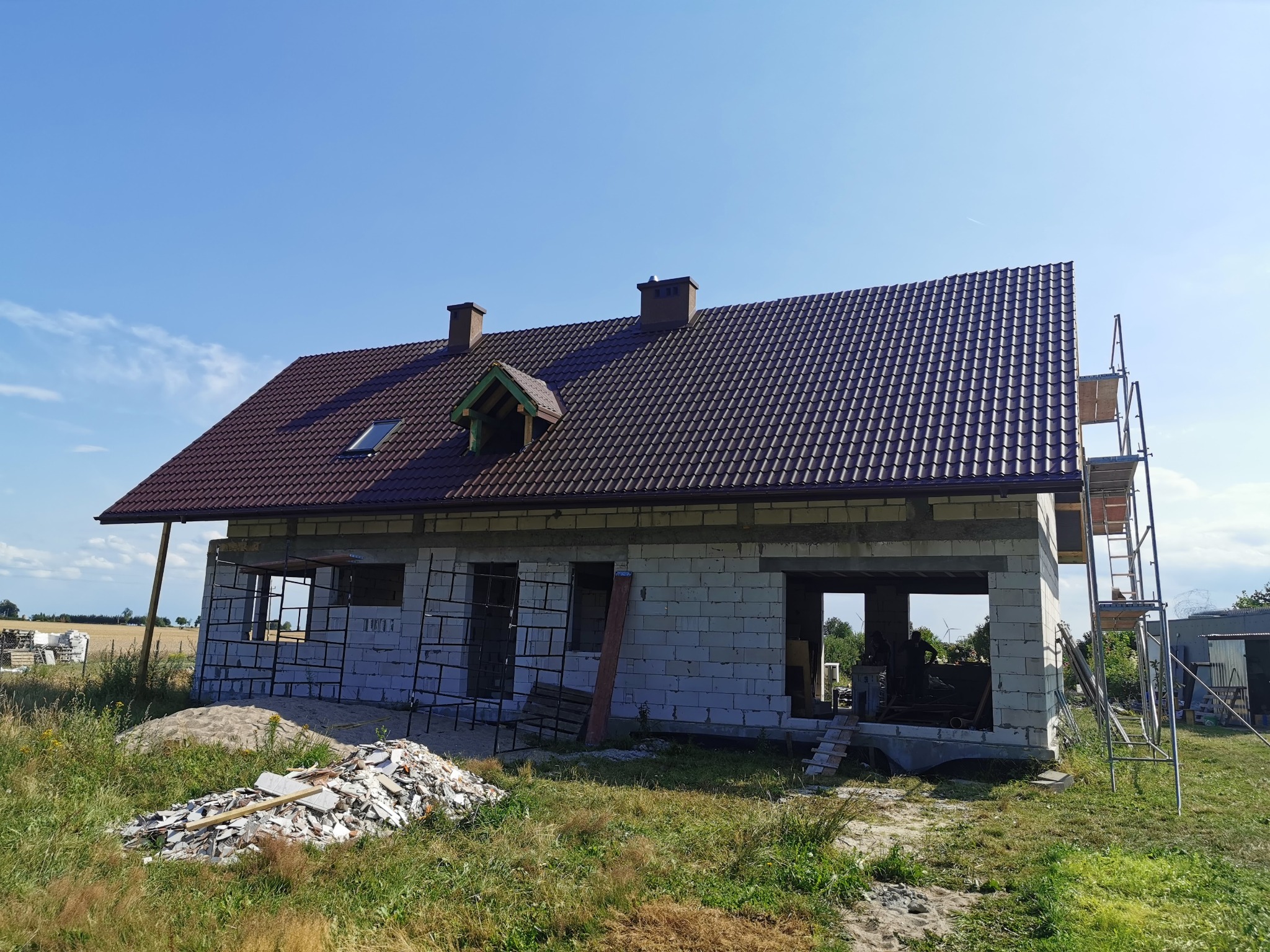 Niedokończony, parterowy budynek z brązowym dachem z dachówki, widoczne rusztowanie, okno dachowe i sterta gruzu przed budynkiem, w tle pola uprawne.