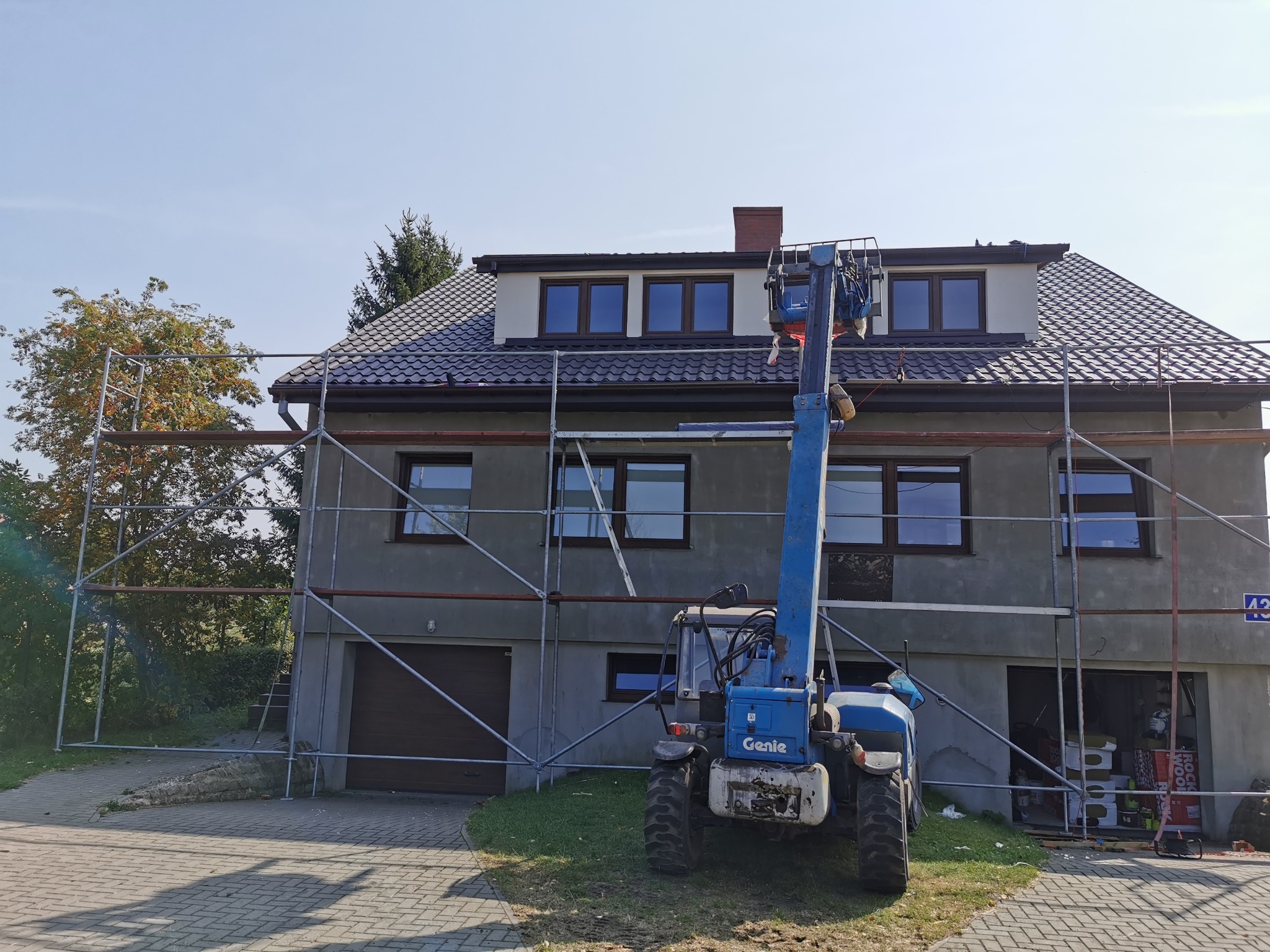 Murowany dom w trakcie remontu elewacji, otoczony rusztowaniem i podnośnikiem Genie, widoczne materiały budowlane w garażu.