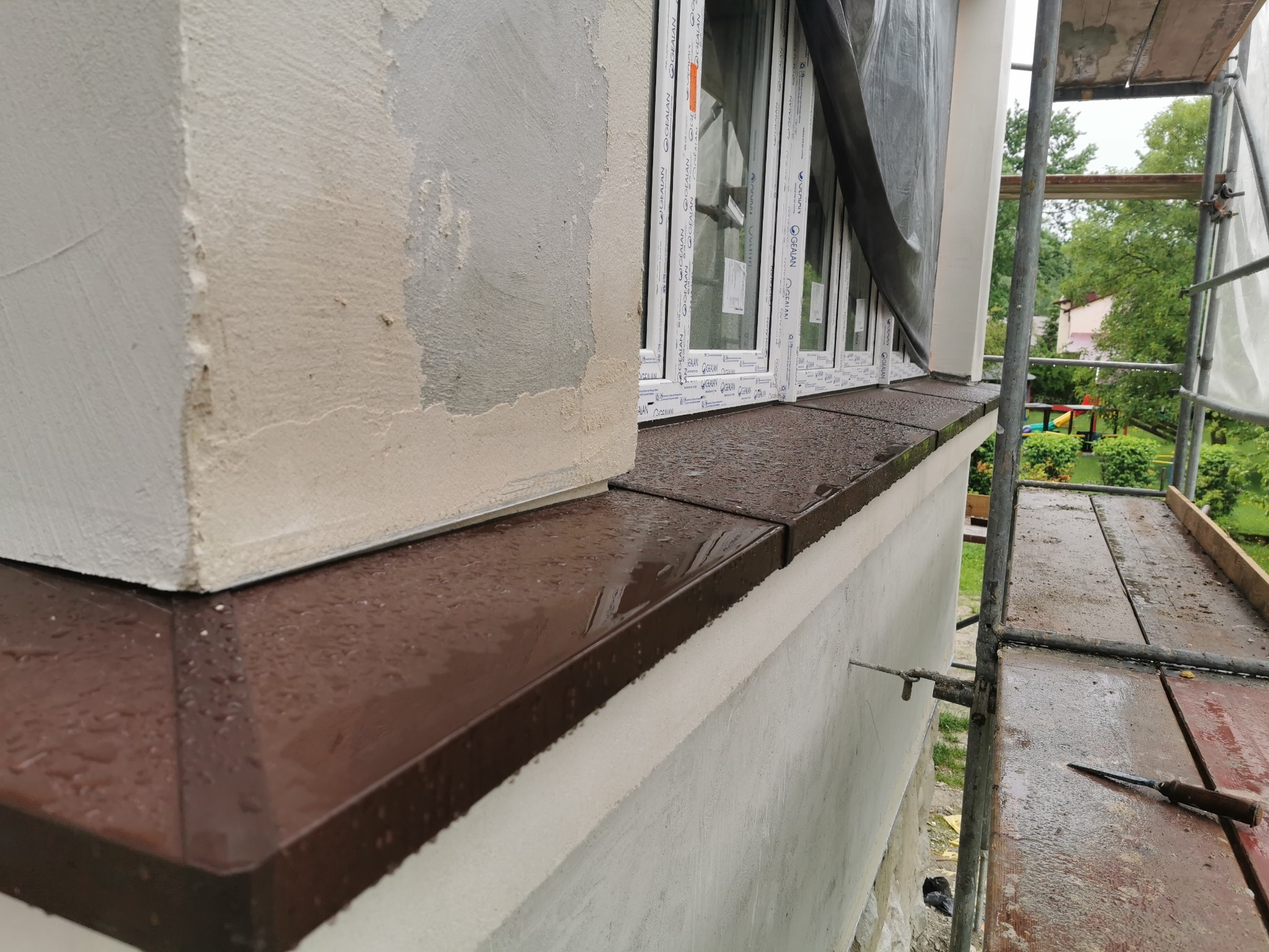 Montaż brązowych, blaszanych parapetów zewnętrznych pod oknami z białymi ramami, widoczne ślady świeżego tynku na elewacji i rusztowanie obok.