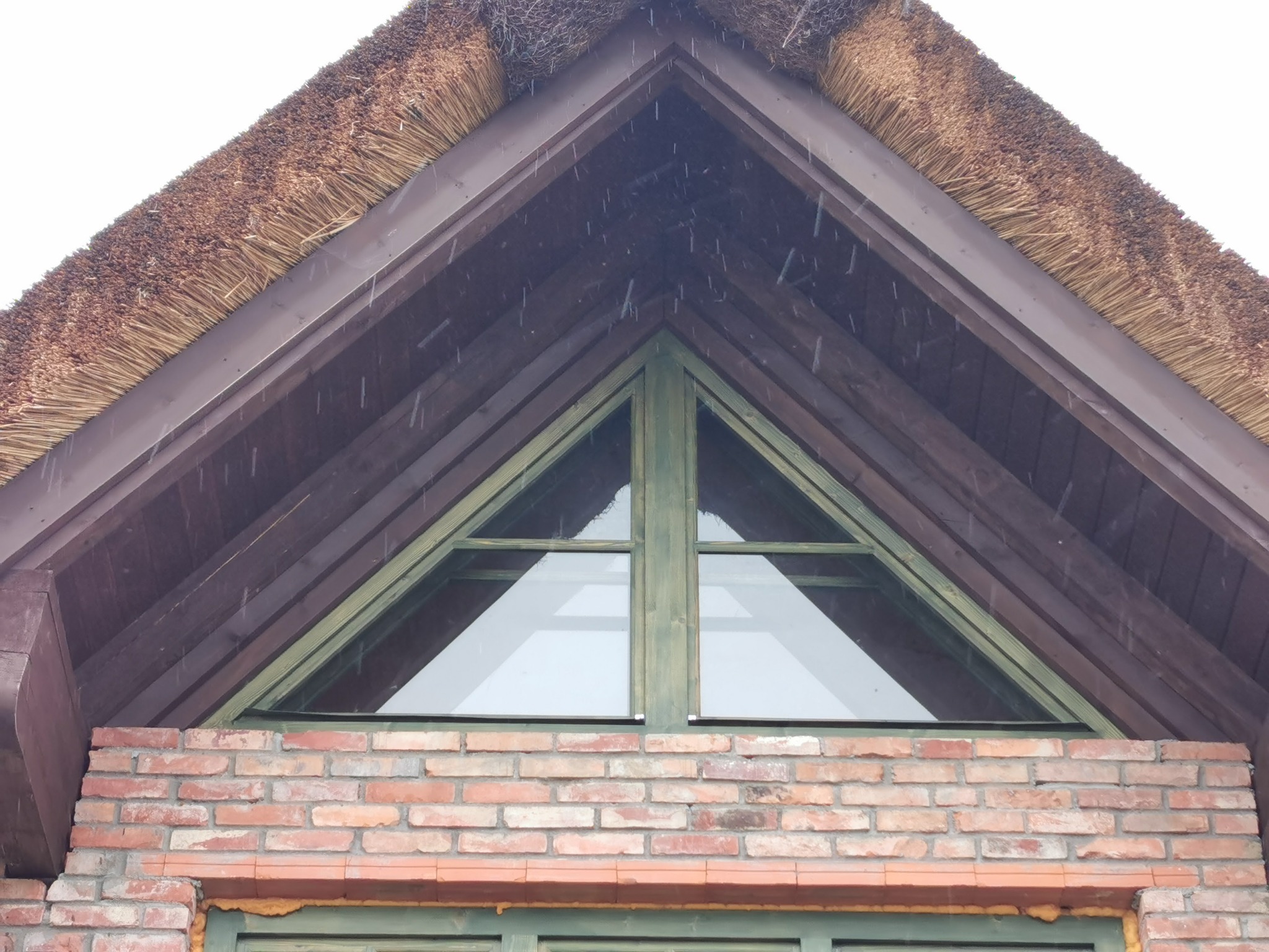 Fragment domu z dachem krytym strzechą, trójkątne okno z zieloną ramą nad murem z czerwonej cegły, widoczne drewniane elementy konstrukcyjne dachu.