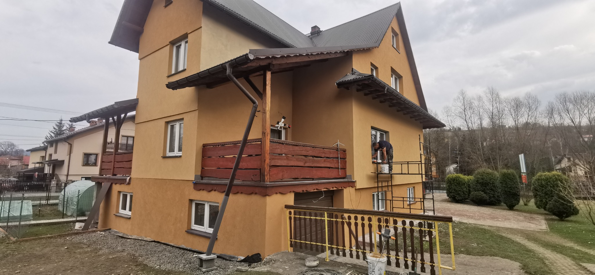 Pracownik na rusztowaniu maluje elewację dwupiętrowego domu z drewnianymi balkonami, widoczne narzędzia i puszki z farbą.