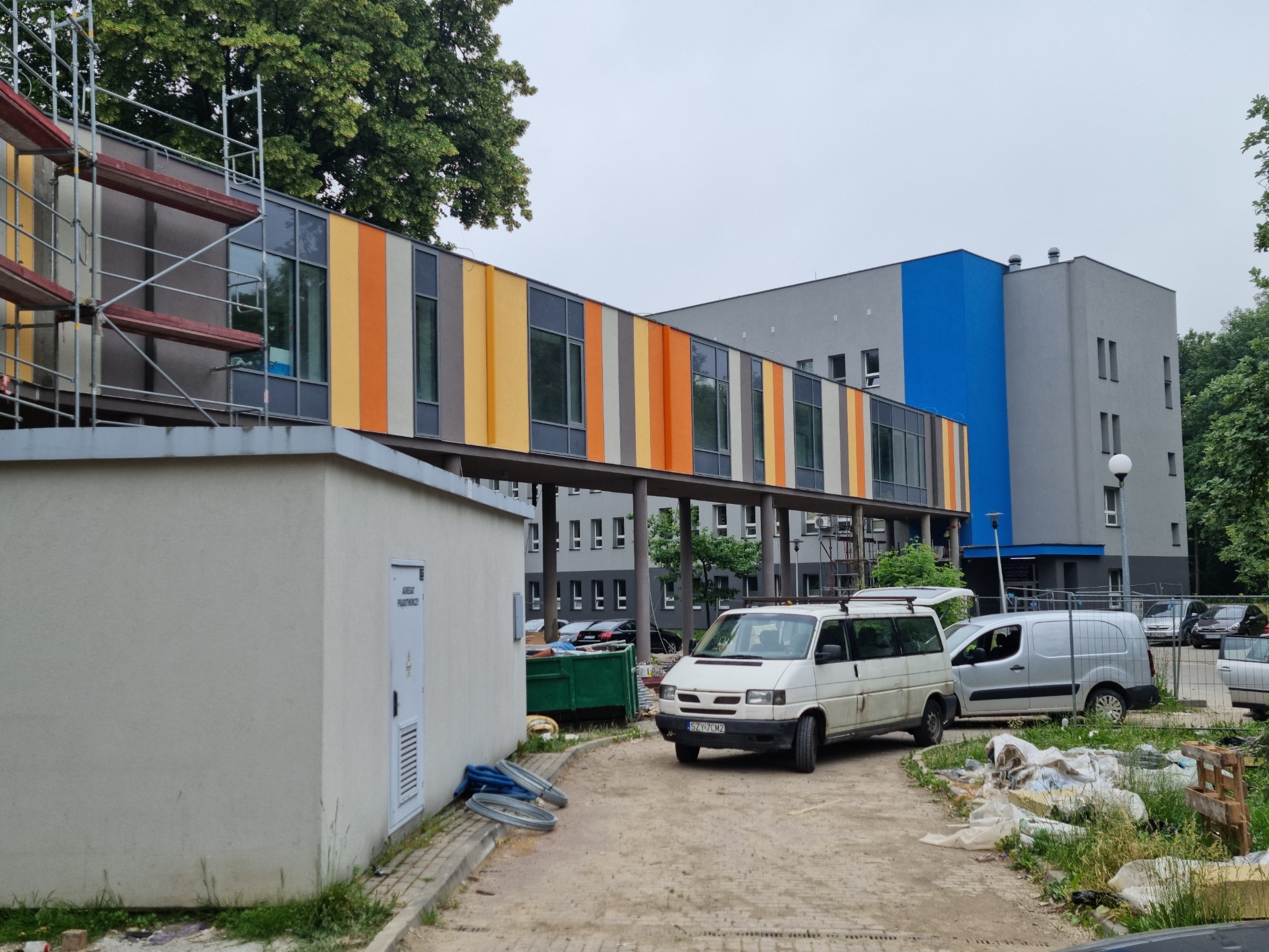 Wielopiętrowy budynek z kolorowymi panelami elewacyjnymi w odcieniach żółci i pomarańczy, widoczne rusztowania, zaparkowane busy i elementy placu budowy.