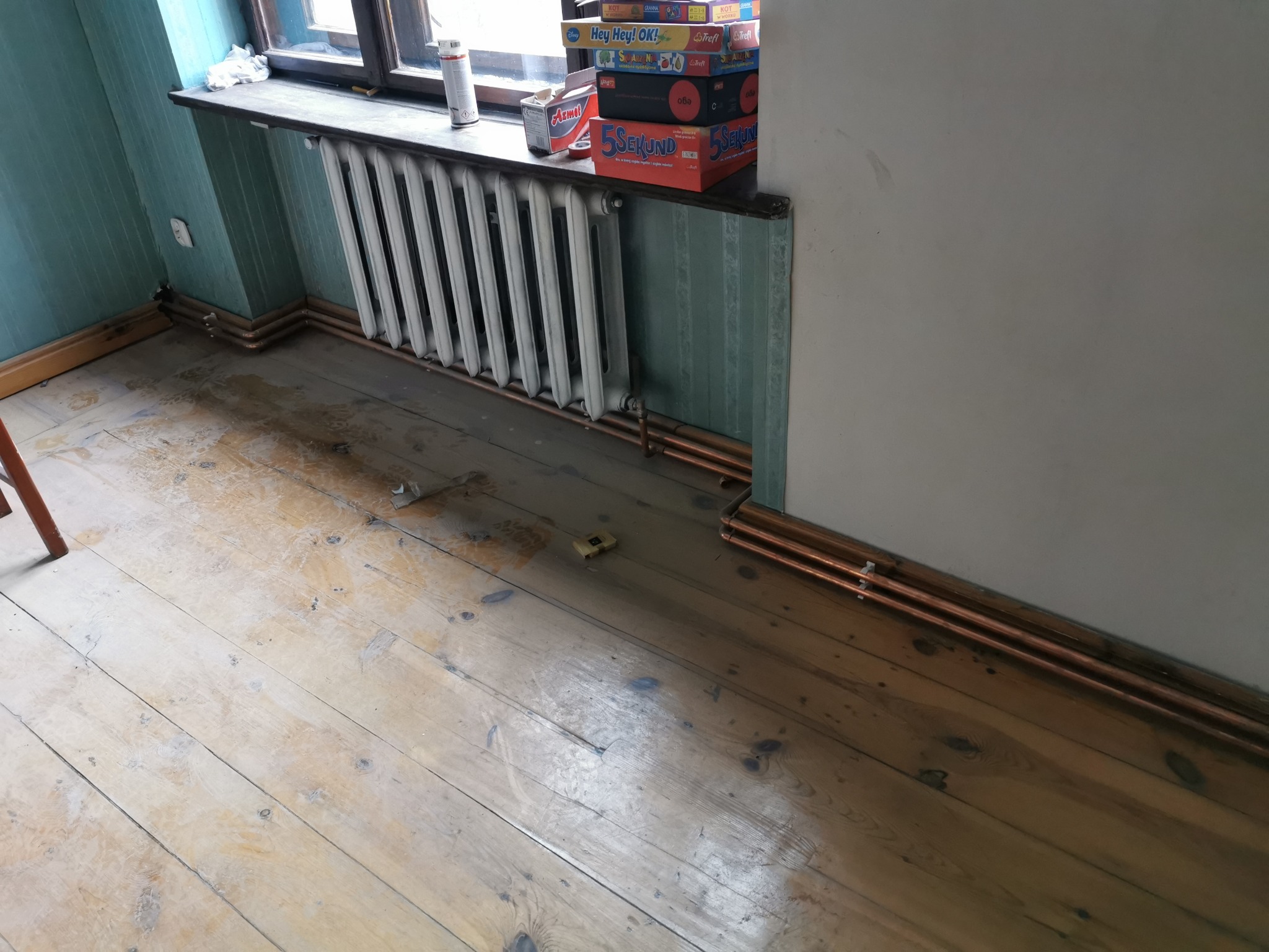 Pokój z drewnianą podłogą, grzejnikiem żeberkowym pod oknem i rurami miedzianymi wzdłuż ściany. Na parapecie stoją gry planszowe i opakowania po produktach. Widoczny fragment krzesła po lewej stronie.