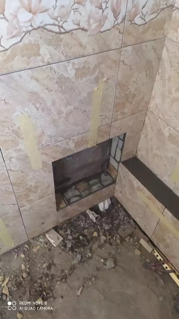 Wnętrze łazienki w trakcie remontu, widoczne płytki ścienne imitujące marmur z motywem kwiatowym, wycięty otwór w ścianie, fragment posadzki z gruzem i resztkami materiałów budowlanych, taśma...